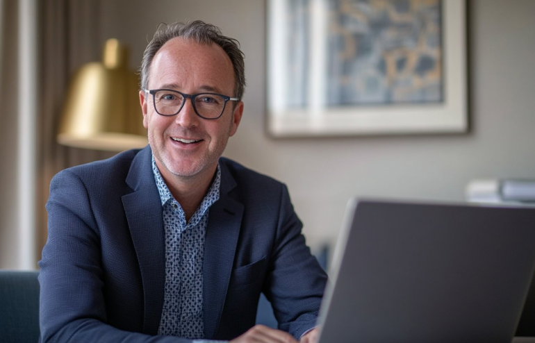 Smiling man in a navy blazer and glasses sits at a desk with a laptop in a bright office.