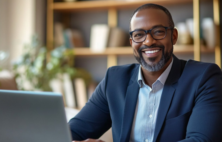 Smiling man with glasses in a navy blazer sits at a desk with a laptop in a bright home office.